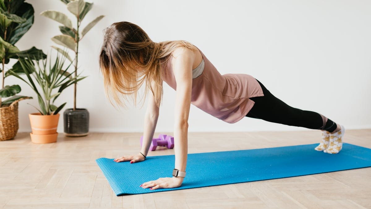 Side view of slender anonymous blond female in sports clothes standing in Plank pose on yoga mat near potted plants on floor at home in daylight